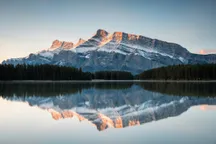 A snow-streaked mountain ridge lit orange by low sun, mirrored in still water below a dark treeline.
