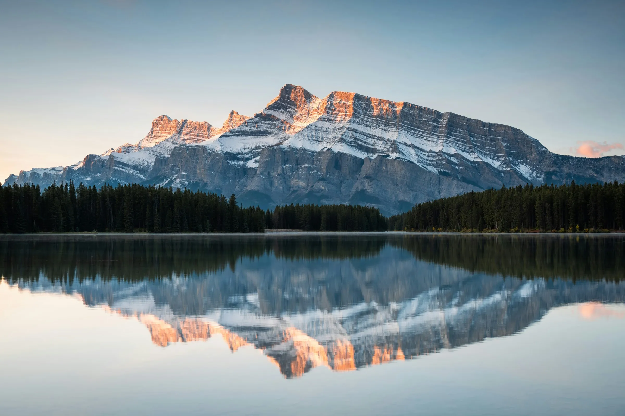 A snow-streaked mountain ridge lit orange by low sun, mirrored in still water below a dark treeline.